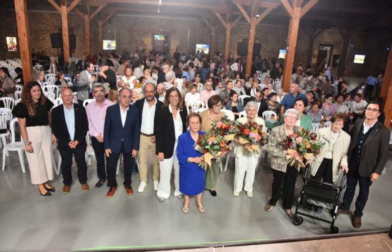 Grupo posa junto a varias mujeres mayores con ramos, ante un público sentado en un salón.