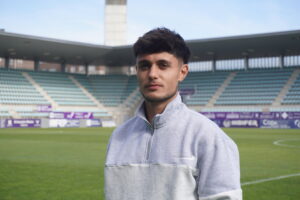 Hombre posa en primer plano sobre el césped de un estadio de fútbol.