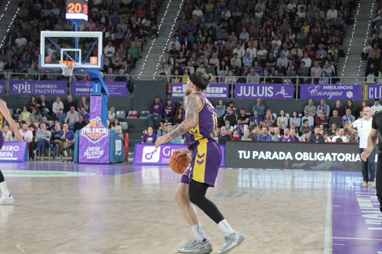 Jugador de baloncesto con uniforme morado y amarillo sostiene el balón ante la canasta.