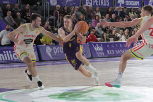 Jugador de baloncesto avanza con el balón mientras dos rivales intentan defenderlo.