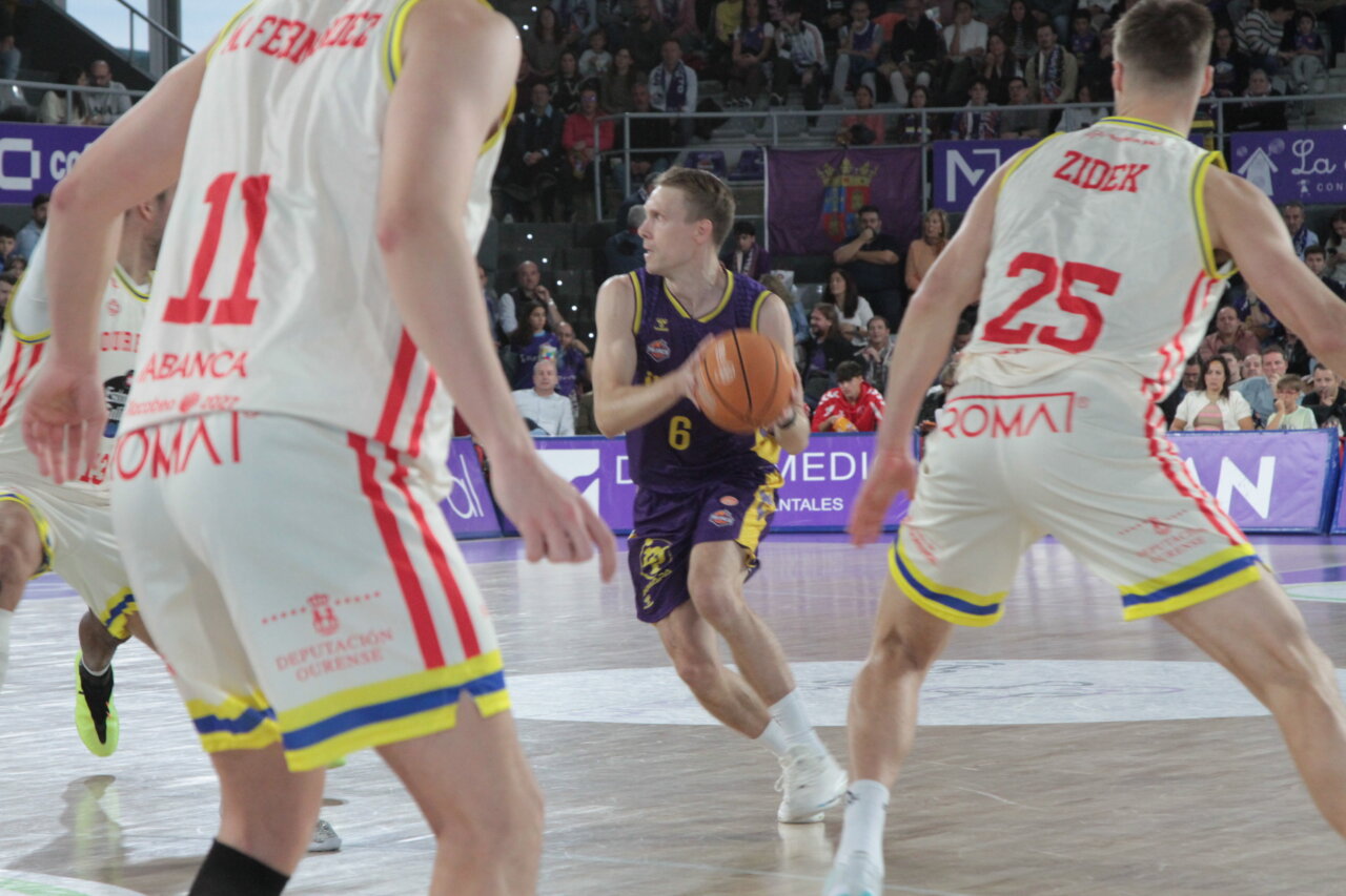 Jugador de baloncesto con el balón, rodeado por dos rivales durante una jugada.