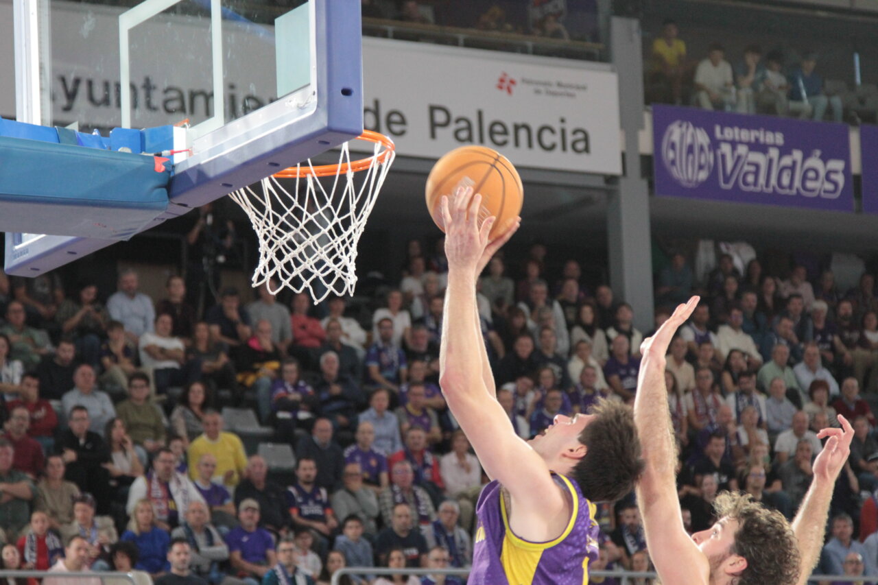Jugador de baloncesto lanza el balón hacia la canasta ante otro jugador y público