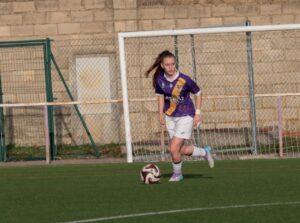 Futbolista con camiseta morada avanza con el balón frente a una portería.