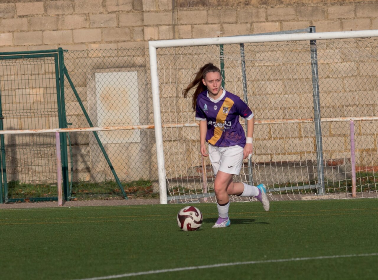 Futbolista con camiseta morada avanza con el balón frente a una portería.