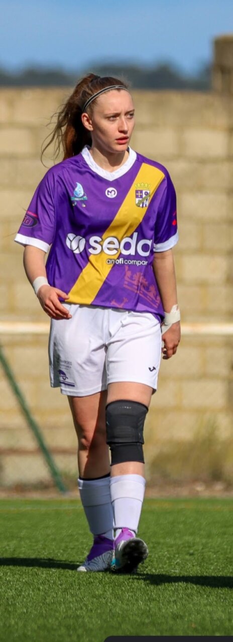 Futbolista con uniforme morado y amarillo camina sobre el césped de una cancha.