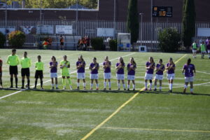 Jugadoras con camisetas moradas forman una fila junto a árbitros en un campo de fútbol