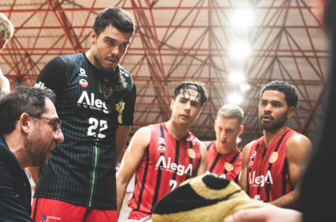 Jugadores de baloncesto escuchan al entrenador mientras señala una pizarra táctica.