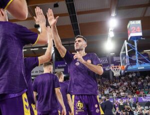 Dos jugadores de baloncesto con camisetas moradas chocan las manos junto a la pista.