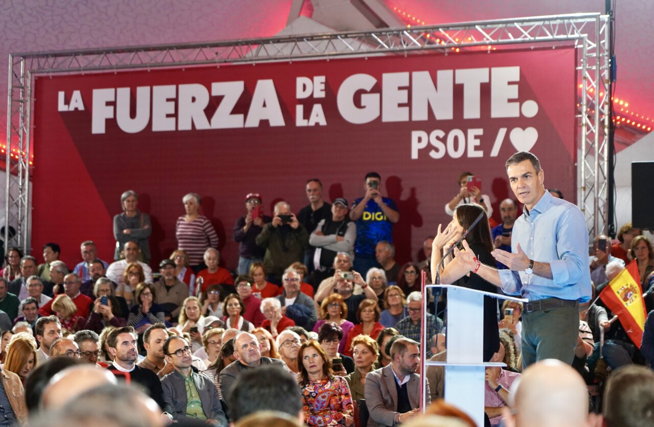 Pedro Sánchez habla ante un público bajo una pancarta roja del PSOE en un mitin.