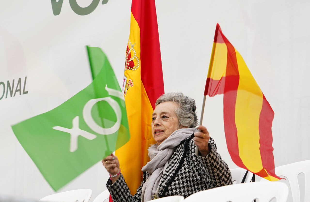 Una mujer sostiene banderas de Vox y de España frente a un fondo con el logotipo.