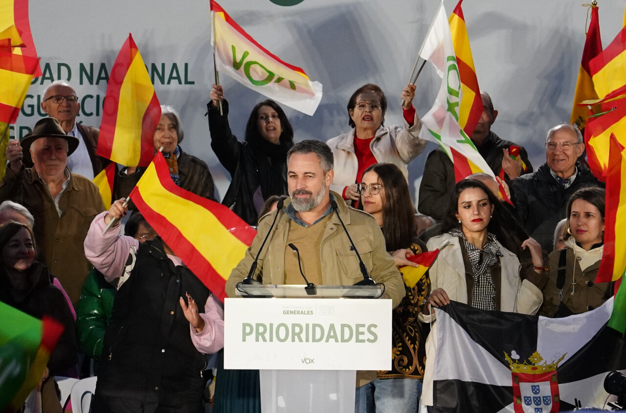 Un orador ante un atril de VOX, rodeado de asistentes con banderas españolas.