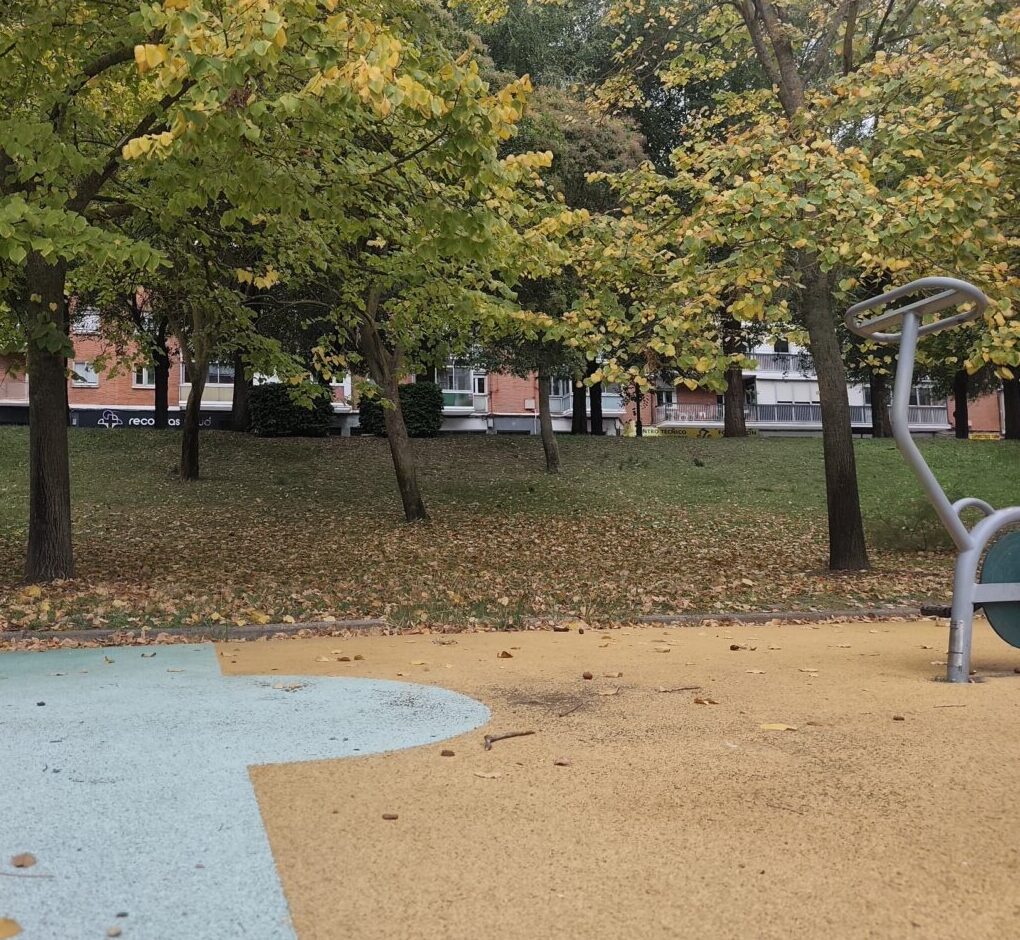 Parque arbolado con suelo de juegos de colores y parte de un aparato