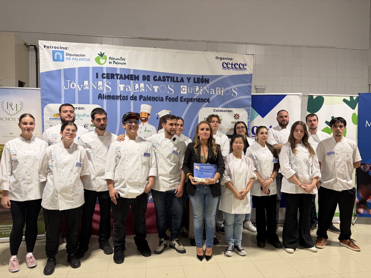 Grupo de participantes con chaquetas blancas posa ante el cartel del certamen culinario.