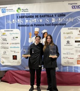 Dos mujeres con chaquetas de cocina sostienen placas ante un cartel del certamen gastronómico.