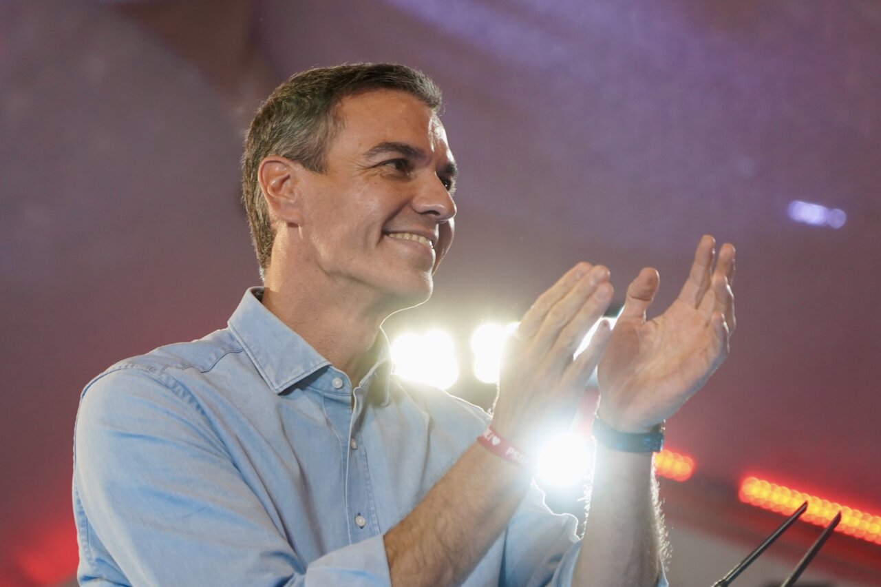 Pedro Sánchez aplaude bajo focos durante un acto político, con camisa azul clara.