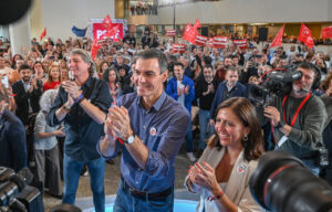 Pedro Sánchez aplaude junto a una mujer mientras una multitud sostiene banderas rojas.