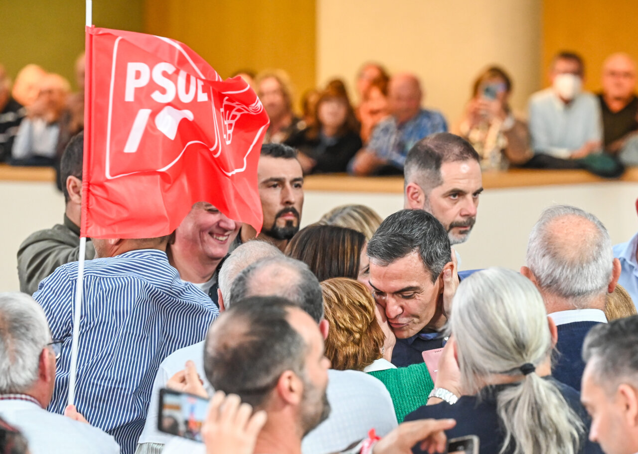 Pedro Sánchez entre asistentes mientras una bandera roja del PSOE ocupa el primer plano.