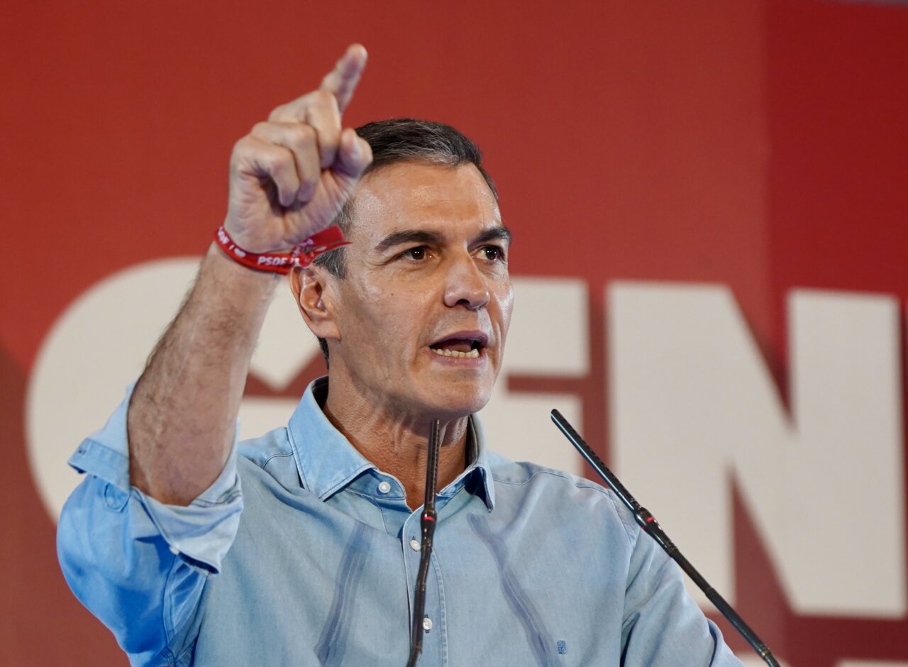 Pedro Sánchez habla ante micrófonos y levanta un dedo frente a un fondo rojo.