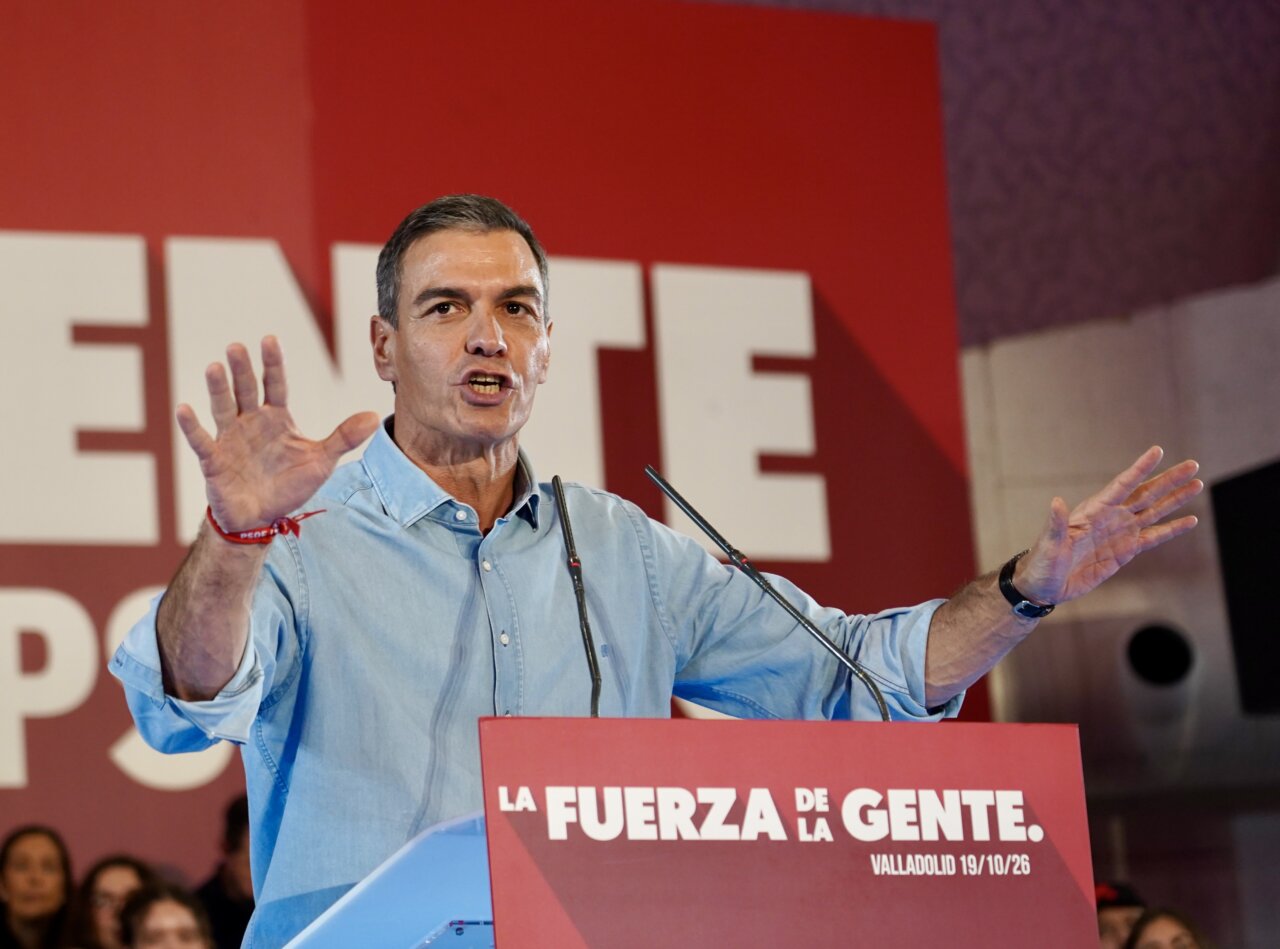 Pedro Sánchez habla ante un atril rojo, con los brazos levantados y público al fondo