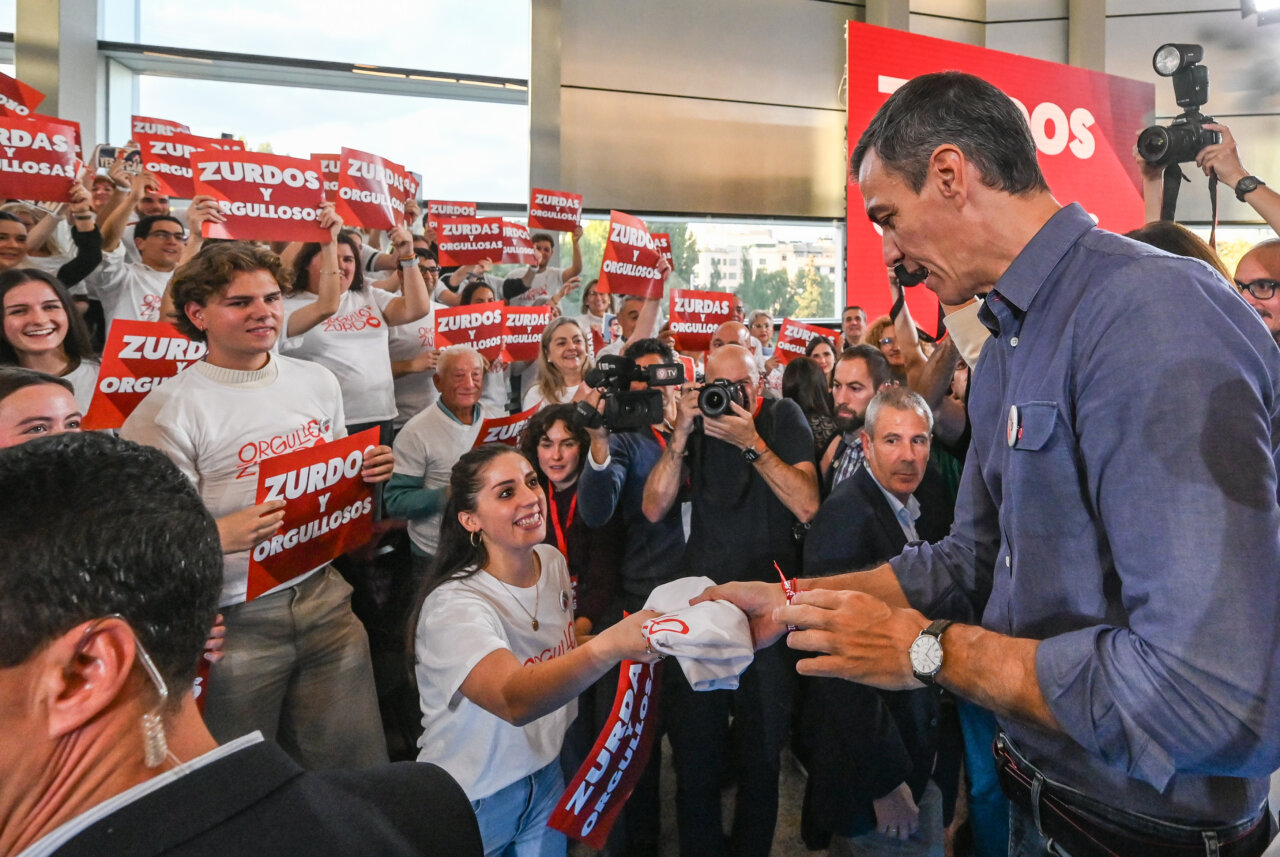 Pedro Sánchez recibe una prenda blanca mientras asistentes levantan carteles de orgullo zurdo