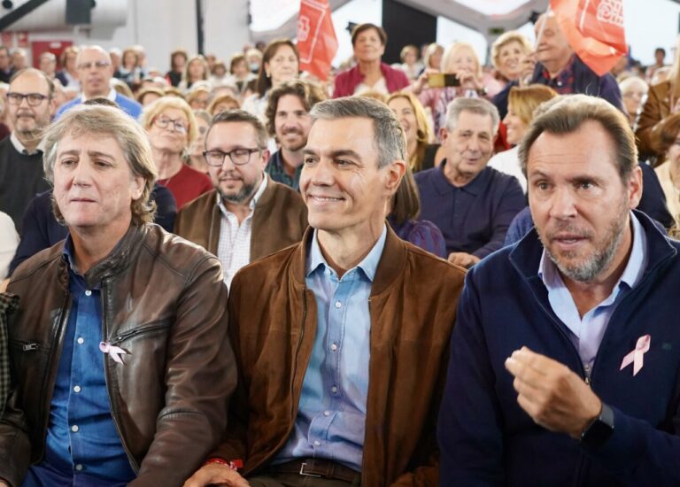 Pedro Sánchez sentado junto a otros asistentes, con banderas rojas al fondo.