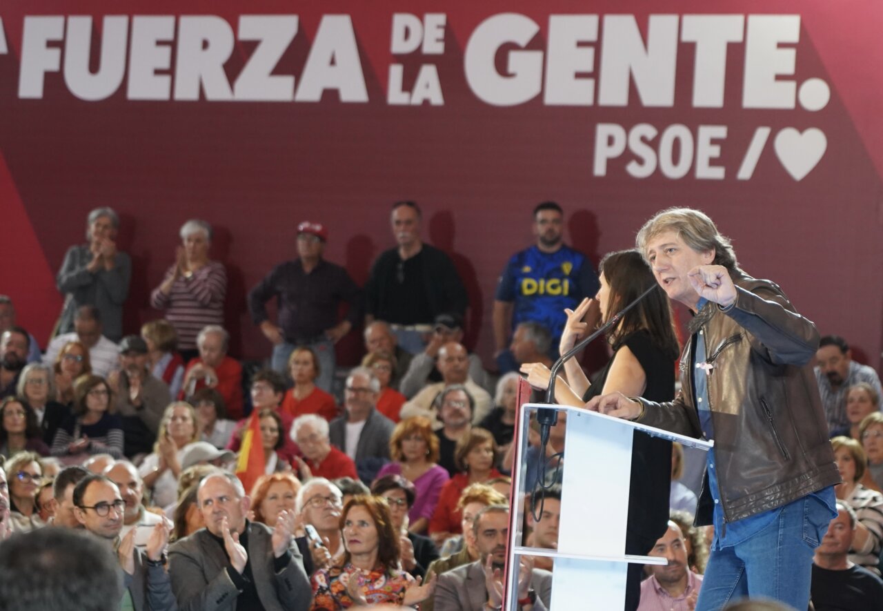Un hombre gesticula junto al atril ante un público sentado y una pancarta del PSOE