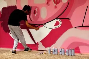 Persona pinta con aerosol un mural rosa mientras varios botes descansan en el suelo.