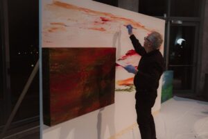 Una persona pinta trazos naranjas sobre un gran lienzo junto a otra obra abstracta.