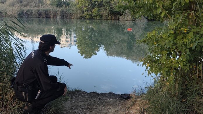 Una persona señala el agua desde la orilla, junto a una boya roja flotante.
