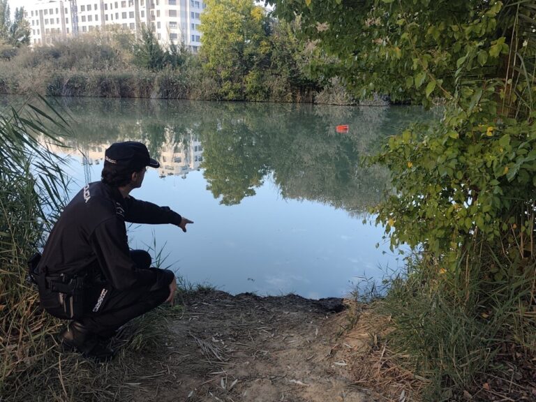 Una persona señala el agua desde la orilla, junto a una boya roja flotante.