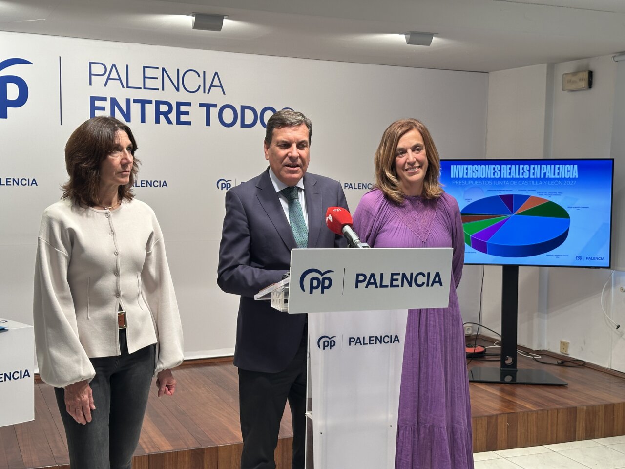Tres personas junto a un atril del PP en Palencia, con un gráfico al fondo.