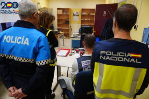 Varias personas con chalecos policiales observan un escritorio y un ordenador en una oficina.