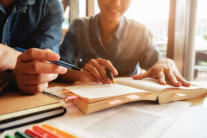Dos personas estudian juntas con libros abiertos y bolígrafos sobre la mesa.