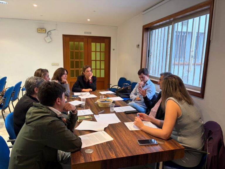 Varias personas conversan alrededor de una mesa con documentos en una sala.