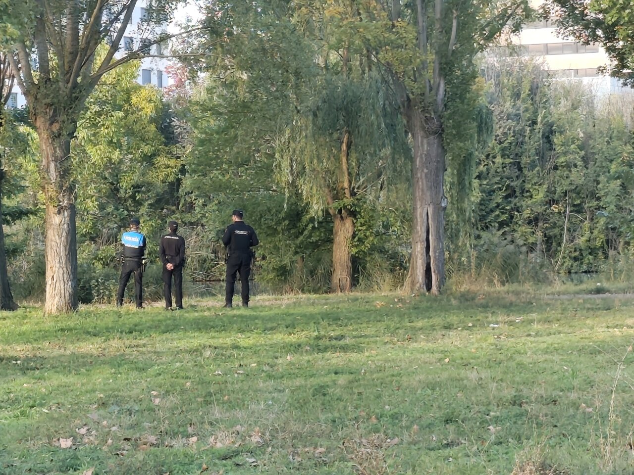Tres personas, una con chaleco azul rotulado Policía, están en una zona arbolada.