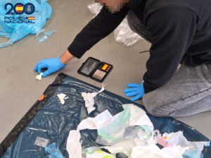 Persona con guantes azules manipula un pequeño paquete junto a una báscula y bolsas de plástico.