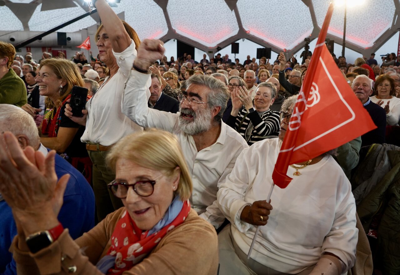 Asistentes aplauden y levantan los brazos entre banderas rojas en un recinto cubierto