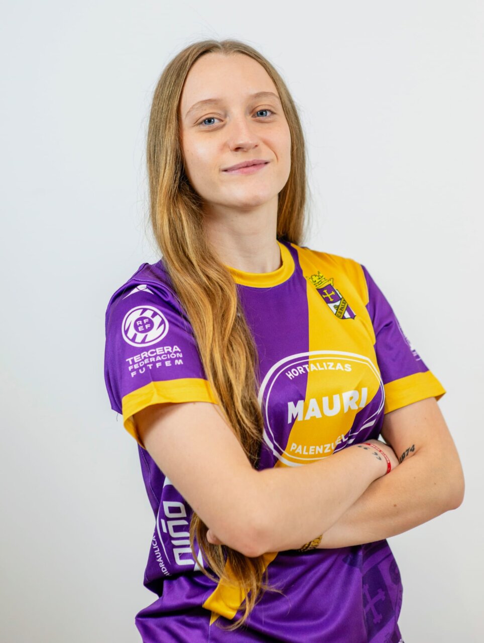 Retrato de una futbolista con camiseta morada y amarilla, brazos cruzados.