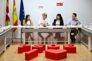 Cuatro personas sentadas en una mesa de reunión, con banderas y letras del PSOE.