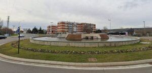Rotonda ajardinada con una fuente circular, edificios residenciales y un autobús al fondo.