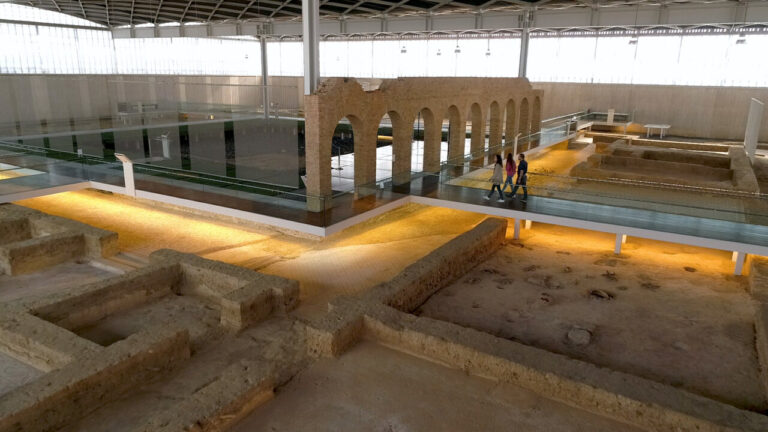 Fotografía de ruinas arqueológicas con arcos de ladrillo y visitantes sobre una pasarela elevada.