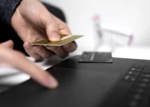 Manos sostienen una tarjeta de pago junto a un ordenador portátil y un carrito desenfocado.