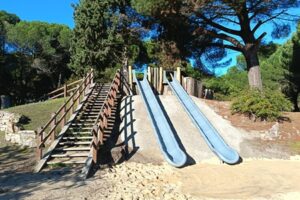 Dos toboganes metálicos junto a una escalera de madera en un parque arbolado.